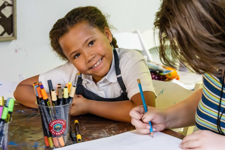 Smiling girl seated at a table with art supplies. 